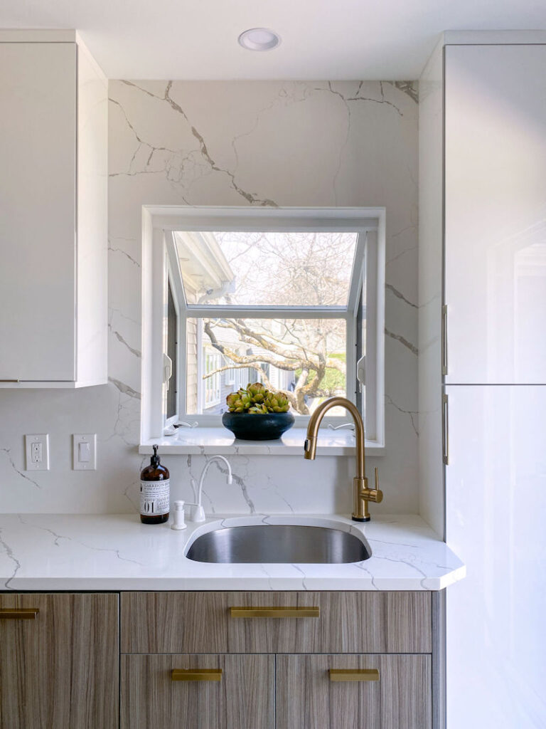 Contemporary kitchen closeup of sink with a brushed gold faucet, full height quartz backsplash, and window box.